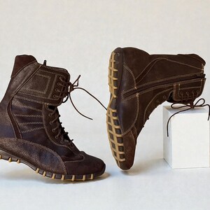 May include: Two brown leather boots with light brown stitching and laces. The boots have a high-top design with a textured sole and are displayed on a white background. One boot is upright, and the other is resting on a white cube.