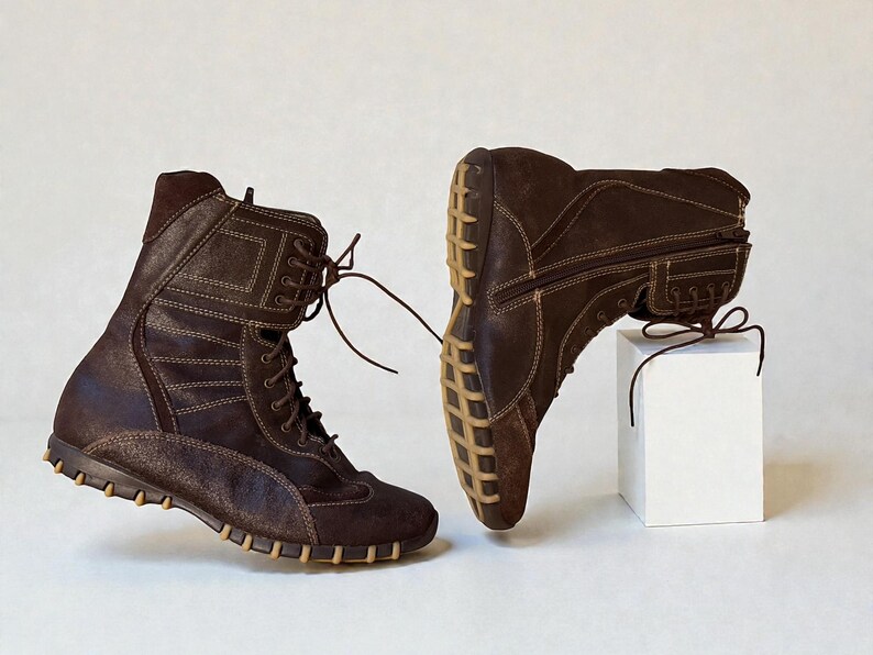 May include: Two brown leather boots with light brown stitching and laces. The boots have a high-top design with a textured sole and are displayed on a white background. One boot is upright, and the other is resting on a white cube.