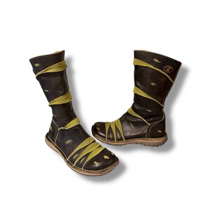 May include: A pair of black leather boots with olive green accents. The boots have a unique design with straps wrapping around the shaft and foot. The soles are a light brown color, and the boots appear to be handmade.