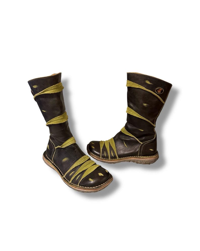 May include: A pair of black leather boots with olive green accents. The boots have a unique design with straps wrapping around the shaft and foot. The soles are a light brown color, and the boots appear to be handmade.