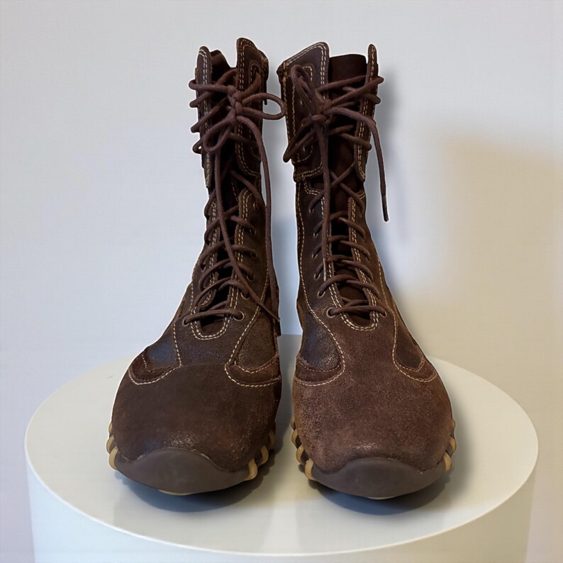 May include: A pair of brown suede lace-up boots. The boots have a high shaft, a rounded toe, and a contrasting light brown sole. The laces are brown and run up the front of the boots. The boots are displayed on a white surface.