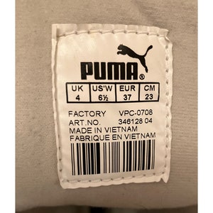 May include: A white Puma shoe label with black text and a black Puma logo. The label shows the shoe size in UK, US, EUR, and CM. The label also includes the factory, art number, and country of origin. The text reads: "PUMA, UK, US'W, EUR, CM, 4, 6 1/2, 37, 23, FACTORY, VPC-0708, ART.NO. 346128 04, MADE IN VIETNAM, FABRIQUE EN VIETNAM."