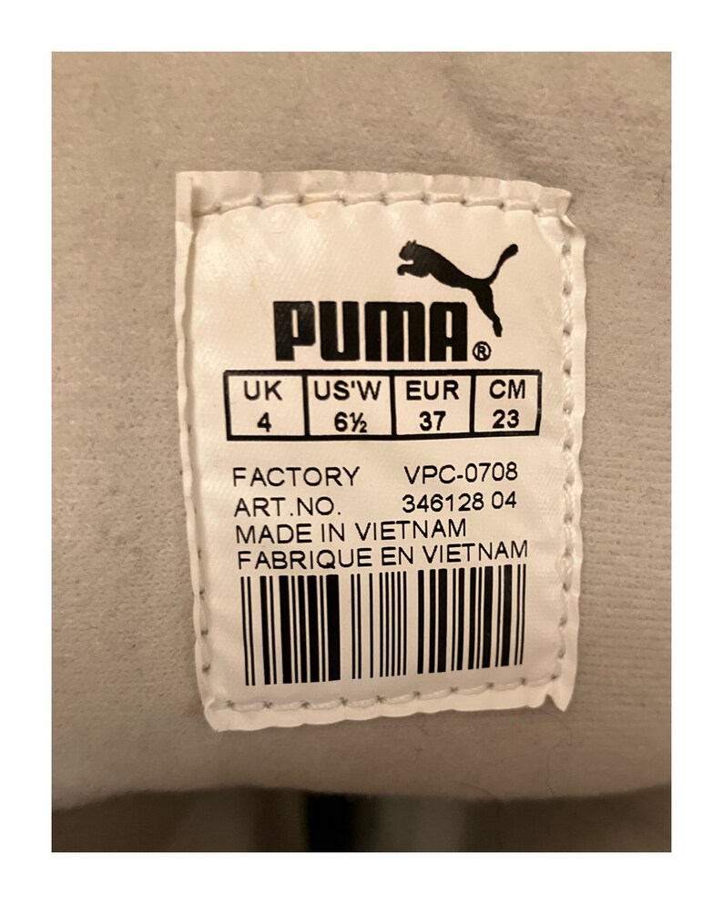 May include: A white Puma shoe label with black text and a black Puma logo. The label shows the shoe size in UK, US, EUR, and CM. The label also includes the factory, art number, and country of origin. The text reads: "PUMA, UK, US'W, EUR, CM, 4, 6 1/2, 37, 23, FACTORY, VPC-0708, ART.NO. 346128 04, MADE IN VIETNAM, FABRIQUE EN VIETNAM."