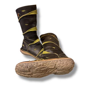 May include: A pair of black leather boots with lime green accents. The boots have a unique, textured sole and a decorative emblem on the side. The design features a folded-over top and stitched detailing, creating a distinctive, handcrafted look.