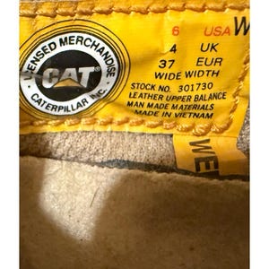 May include: Close-up of a yellow tag with the text "LICENSED MERCHANDISE CAT CATERPILLAR INC." and size information: USA 6, UK 4, EUR 37. The tag also states "WIDE WIDTH" and "LEATHER UPPER BALANCE MAN MADE MATERIALS MADE IN VIETNAM."