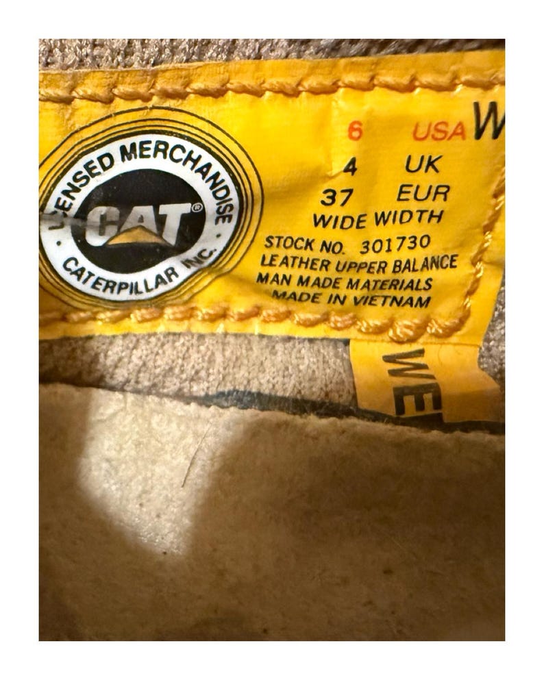 May include: Close-up of a yellow tag with the text "LICENSED MERCHANDISE CAT CATERPILLAR INC." and size information: USA 6, UK 4, EUR 37. The tag also states "WIDE WIDTH" and "LEATHER UPPER BALANCE MAN MADE MATERIALS MADE IN VIETNAM."