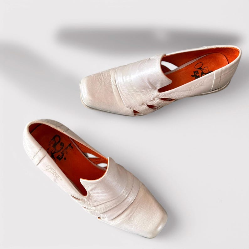 May include: A pair of white leather shoes with cutouts and an orange insole. The shoes have a square toe and a low heel. The brand name "Fly" is visible on the insole.