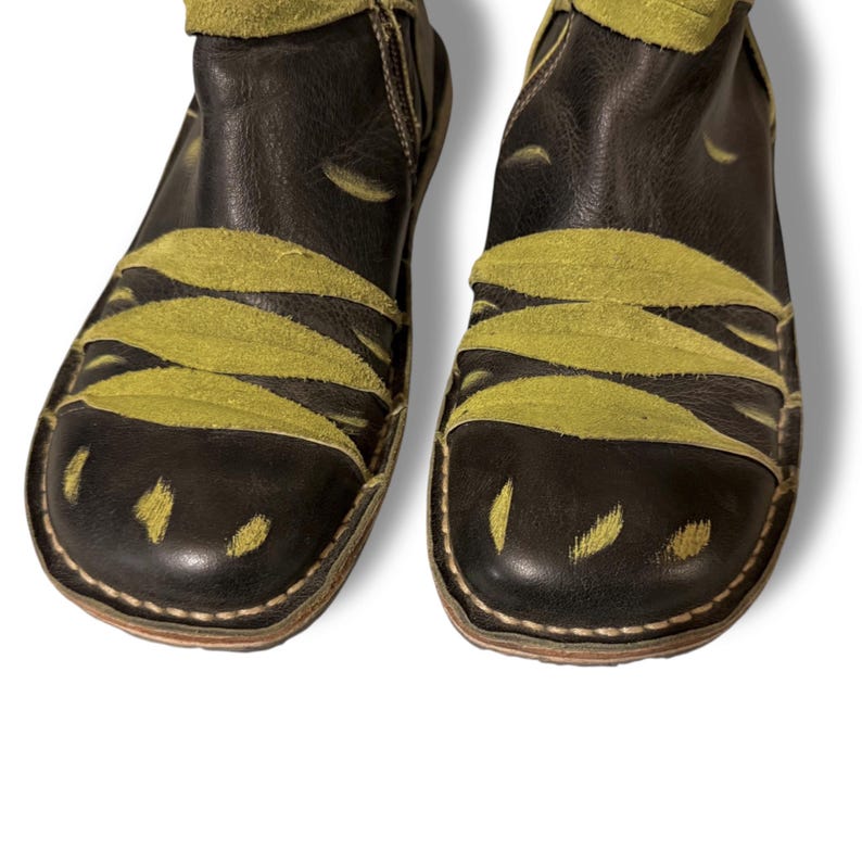 May include: A pair of black leather ankle boots with olive green suede accents. The boots feature a unique design with curved suede overlays and a stitched sole. The boots have a side zipper and a rounded toe.