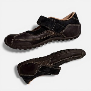 May include: Two brown leather shoes with a velcro strap closure. The shoes have a textured sole and white stitching details. The upper part of the shoe is made of a combination of leather and suede.