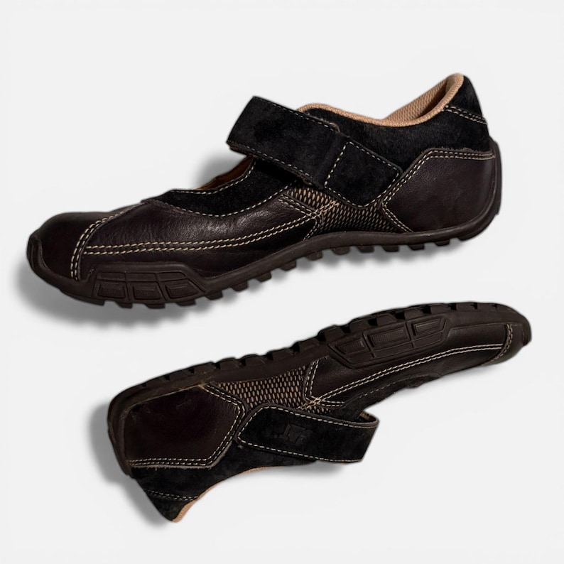 May include: Two brown leather shoes with a velcro strap closure. The shoes have a textured sole and white stitching details. The upper part of the shoe is made of a combination of leather and suede.