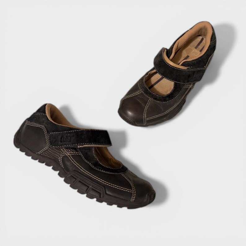 May include: Two black leather shoes with a Mary Jane style, featuring a velcro strap and white stitching. The shoes have a textured sole and a suede-like accent on the top. The shoes are displayed on a white background.