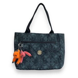 Peut inclure: Sac fourre-tout teal avec un motif floral noir et des poignées noires. Il possède une poche zippée et un logo rond. Un porte-clés avec deux breloques en peluche, une orange et une rose, est attaché.