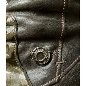 May include: Close-up of a brown leather boot with a decorative metal button. The boot has a contrasting green leather panel.