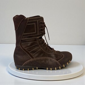 May include: A brown suede boot with a high top and a lace-up design. The boot features a textured sole and decorative stitching. The boot is displayed on a white surface.