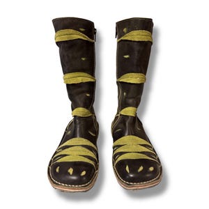 May include: A pair of dark brown leather boots with olive green suede accents. The boots have a knee-high design with a zipper closure and decorative cutouts. The olive green suede is used in bands across the boots.