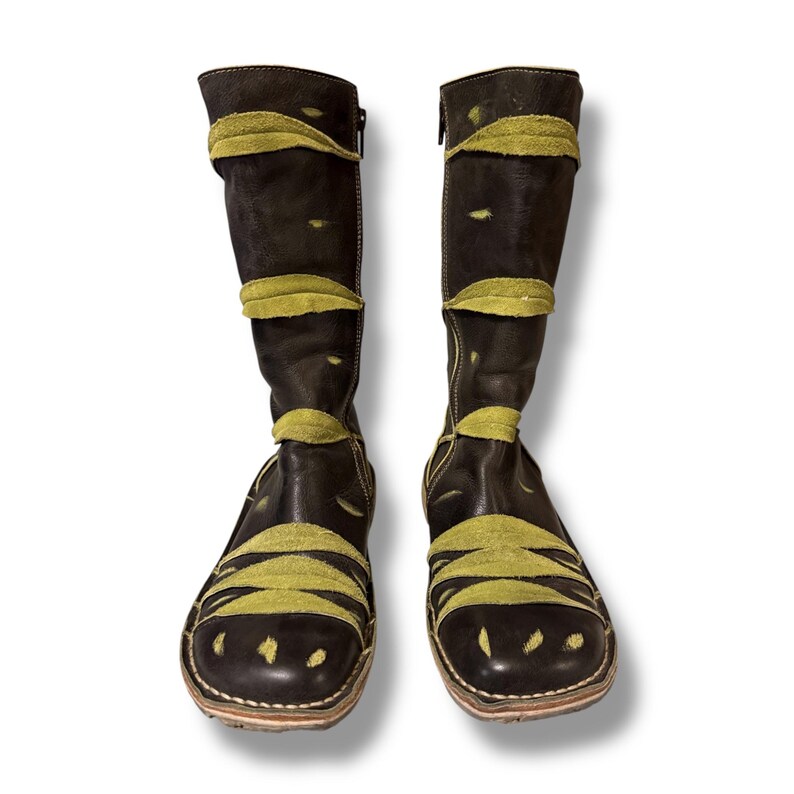 May include: A pair of dark brown leather boots with olive green suede accents. The boots have a knee-high design with a zipper closure and decorative cutouts. The olive green suede is used in bands across the boots.