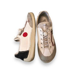 May include: A pair of white leather sneakers with red accents and a zipper detail. The sneakers have a white rubber sole and a black trim around the ankle.