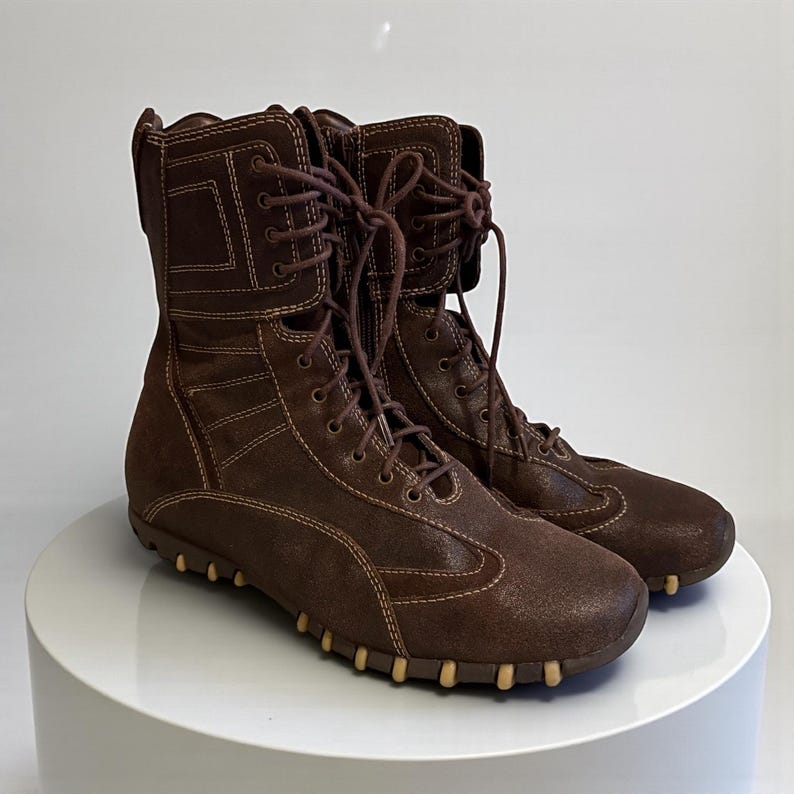 May include: A pair of brown leather boots with a lace-up design. The boots have a mid-calf height, with a textured finish and contrasting stitching. The soles feature small, light-colored pegs. The boots are displayed on a white surface.