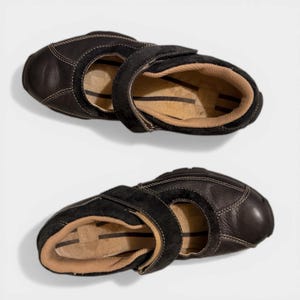 May include: Two pairs of black leather shoes with a strap closure. The shoes have a rounded toe and a tan interior. The stitching is visible in a lighter color, and the shoes appear to be unworn.