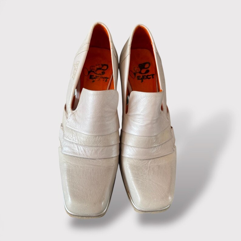 May include: A pair of white leather shoes with a square toe and an open design. The shoes have an orange insole with the word "Effect" written on it.