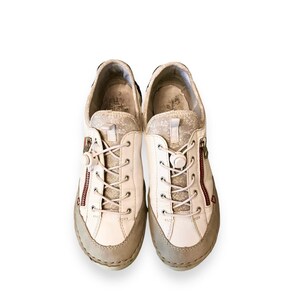 May include: White leather sneakers with a zipper detail on the side. The laces are tied and the shoes have a light brown sole.