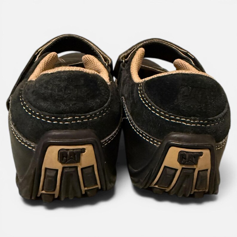 May include: Pair of black suede Caterpillar shoes with tan accents. The shoes have a hook-and-loop closure and the Caterpillar logo on the heel. The soles are brown and the stitching is white. The shoes are designed for casual wear.