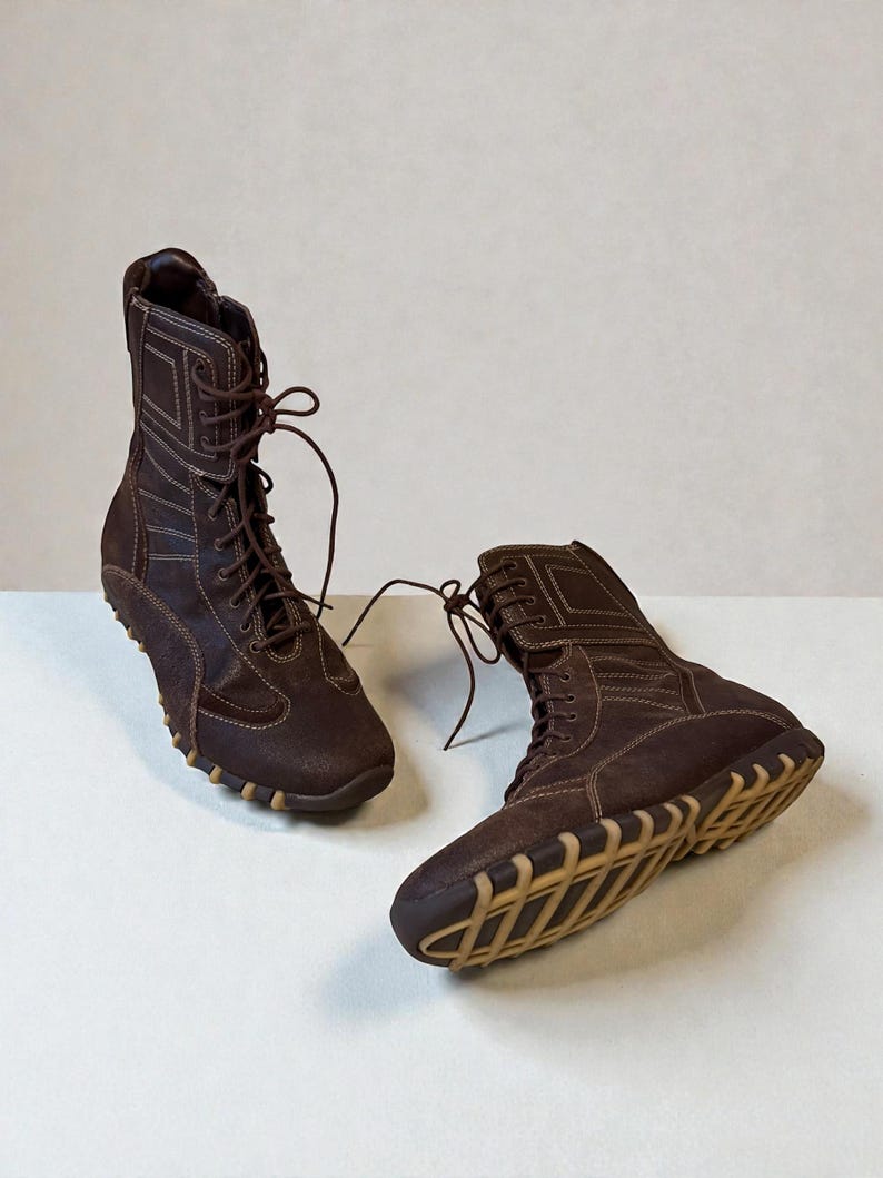 May include: A pair of brown suede boots with a high-top design. The boots feature a lace-up closure, white stitching details, and a tan rubber sole. The boots are designed for casual wear.