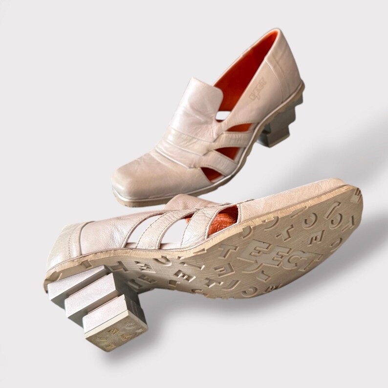 May include: A pair of beige leather heeled shoes with cutouts and a chunky platform heel. The shoes have a unique design with the word "TECT" embossed on the sole.