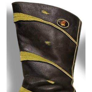 May include: A tall, dark brown leather boot with olive green suede accents. The boot features a decorative design with a gold trim and a small emblem near the top. The boot's design includes a curved top and a textured leather finish.