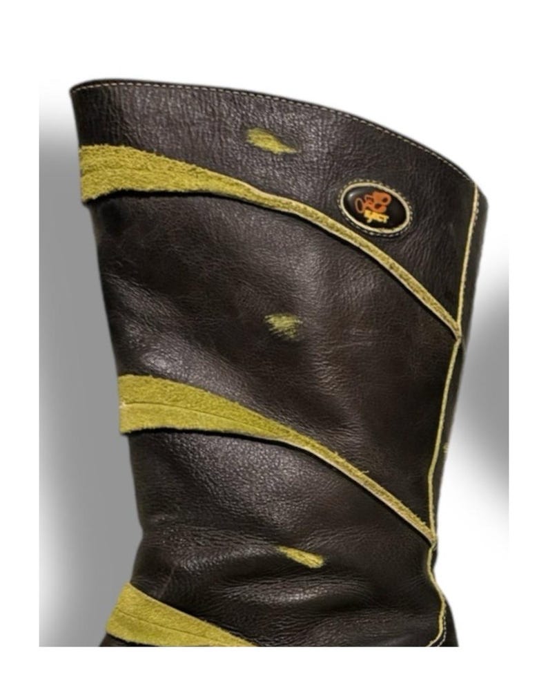May include: A tall, dark brown leather boot with olive green suede accents. The boot features a decorative design with a gold trim and a small emblem near the top. The boot's design includes a curved top and a textured leather finish.