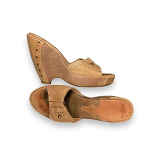 May include: A pair of brown leather wedge sandals with a buckle detail. The sandals have a cork wedge heel and a leather strap across the foot. The brand name "Caswick" is visible on the strap.