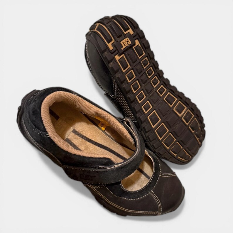 May include: Black leather Mary Jane style shoes with a strap closure. The shoes have a brown sole with the CAT logo and white stitching details. The interior is tan and black striped.