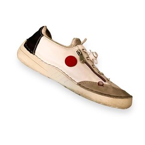 May include: White leather sneaker with a red circle and a zipper detail. The shoe has a white sole and a brown leather heel.