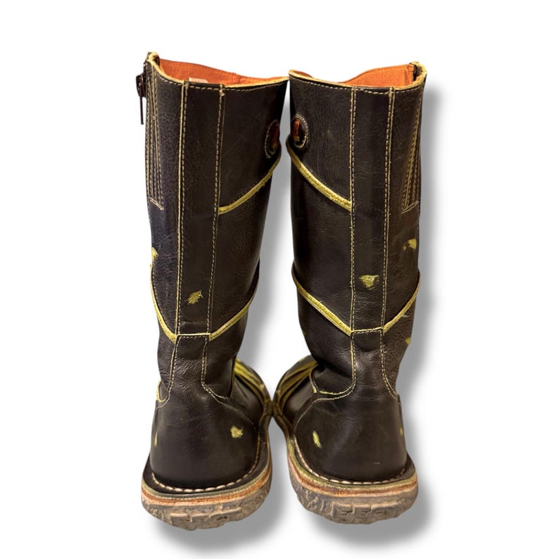 May include: A pair of brown leather boots with yellow stitching and accents. The boots have a zipper on the side and a round emblem near the top. The soles are a light brown color. The boots are knee-high.