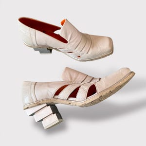 May include: A pair of white leather heeled shoes with a unique block heel design. The shoes have a cut-out design on the upper and a pointed toe.