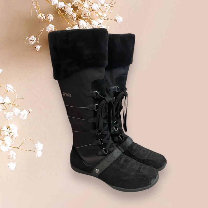 May include: Black knee-high boots with a lace-up front and a faux fur cuff. The boots have a black suede toe and heel. The brand name "Dr. Scholl's" is visible on the side of the boot.