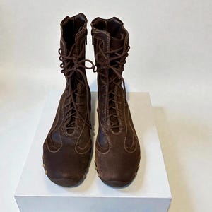 May include: A pair of brown suede and leather boots with laces and zippers. The boots have a round toe and a rubber sole. The boots are displayed on a white surface against a white background.