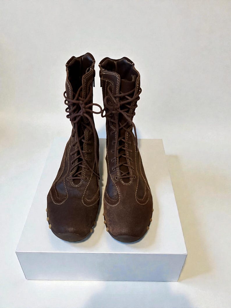 May include: A pair of brown suede and leather boots with laces and zippers. The boots have a round toe and a rubber sole. The boots are displayed on a white surface against a white background.