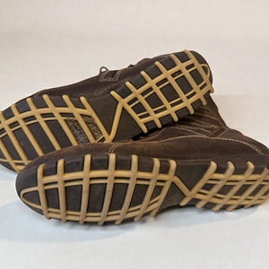May include: Pair of brown leather boots with a light brown grid-patterned sole. The boots have a suede-like texture on the upper part and are laced up. The soles are thick and appear durable, suitable for various terrains.