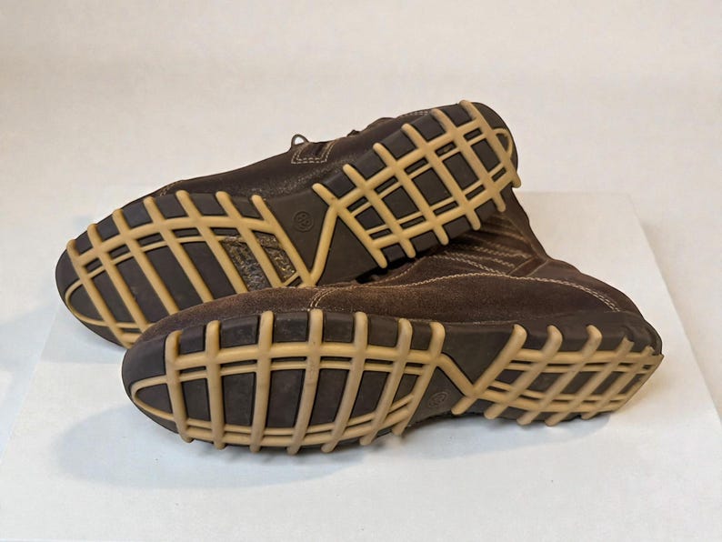 May include: Pair of brown leather boots with a light brown grid-patterned sole. The boots have a suede-like texture on the upper part and are laced up. The soles are thick and appear durable, suitable for various terrains.