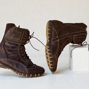May include: Two brown leather boots with light brown stitching and laces. The boots have a high-top design with a textured sole and are displayed on a white background. The boots are a size 9.