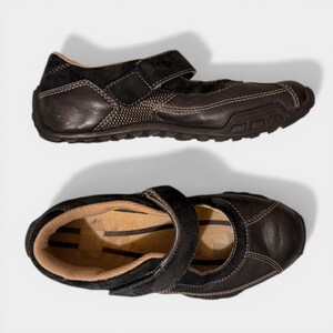 May include: Pair of black leather Mary Jane style shoes with a hook-and-loop strap. The shoes have a textured design on the sides and a brown interior. The soles are dark brown with a treaded pattern.