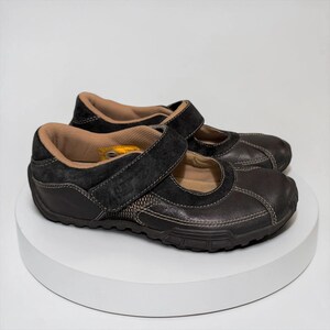 May include: A pair of dark brown Mary Jane style shoes with a hook-and-loop strap closure. The shoes have a black suede accent and tan interior. The soles are dark brown and textured.