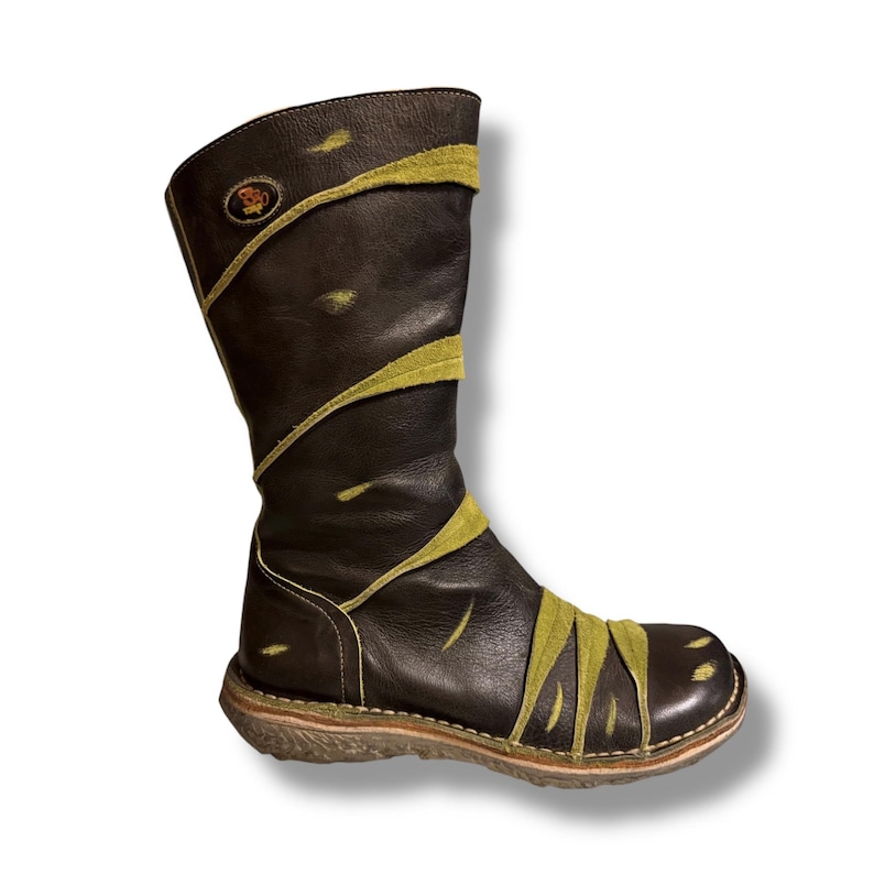 May include: A tall, dark brown leather boot with olive green suede accents. The boot features a round toe, a stitched sole, and a decorative emblem near the top. The suede accents are arranged in a diagonal pattern across the boot.