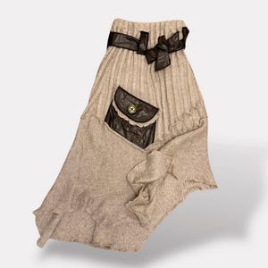 Puede incluir: Falda de punto beige con bolsillo de cuero marrón y lazo decorativo marrón en la cintura. La falda tiene un diseño asimétrico único con un dobladillo con volantes y un detalle de botón en el bolsillo.