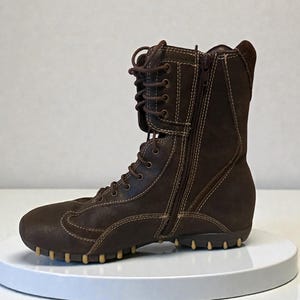 May include: Brown leather boot with a side zipper and lace-up closure. The boot features a rounded toe, a textured sole, and contrasting stitching. The boot is approximately 8 inches tall.