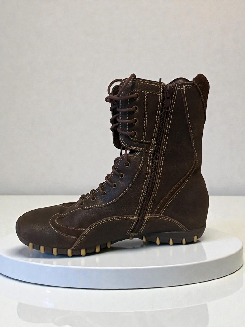 May include: Brown leather boot with a side zipper and lace-up closure. The boot features a rounded toe, a textured sole, and contrasting stitching. The boot is approximately 8 inches tall.