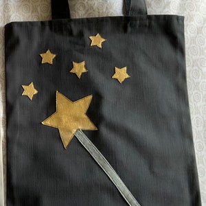 Könnte beinhalten: Schwarze Stofftasche mit einem goldenen Sternenstab und goldenen Sternen, die über die Tasche verteilt sind.