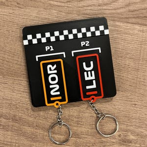 May include: Black rectangular keychain holder with two keychains. One keychain has an orange rectangle with the letters "NOR", the other has a red rectangle with the letters "LEC". Both keychains have silver keyrings.