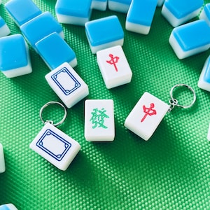 Op de afbeelding: Blauwe en witte Mahjong-tegels op een groene ondergrond. Sommige tegels zijn sleutelhangers. De tegels hebben Chinese karakters in rood en groen. De tegels zijn klein en rechthoekig.